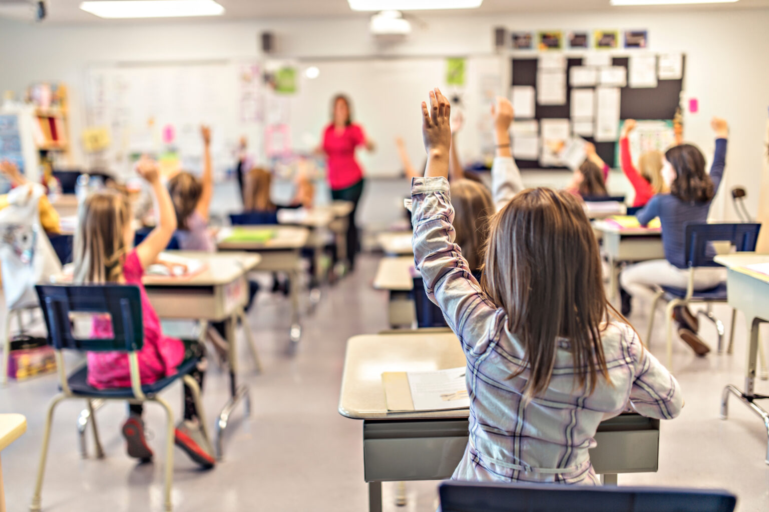 Group of school kids in a classroom. One girl in the foreground with her hand up.