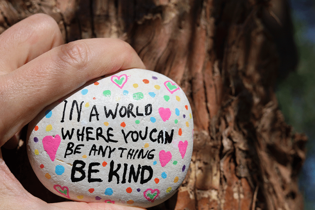A hand holding a painted rock with the words: In a world where you can be anything be kind.