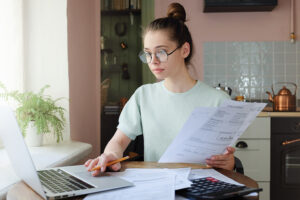 young woman sitting at a kitchen table in front of a laptop, crunching numbers on student loan and student debt paperwork.